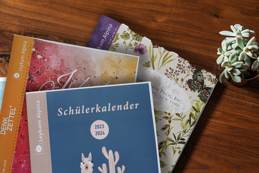 3 Kalender übereinander auf einem Holztisch neben einer Blume, auf dem obersten Kalender steht ,,Schülerkalender 2023, 2024"